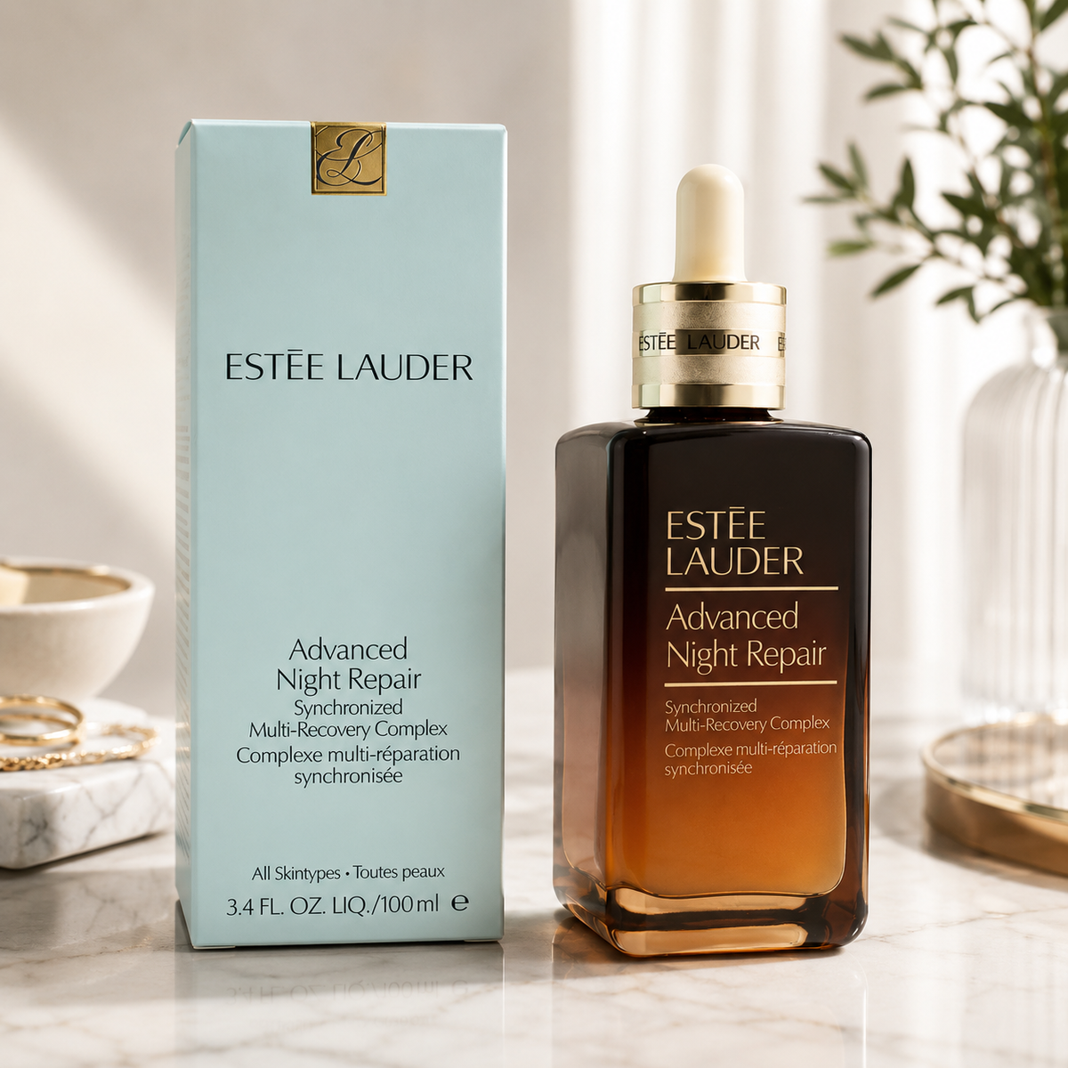Estée Lauder Advanced Night Repair skincare set, featuring a light blue outer box and a dark amber glass dropper bottle with gold lettering, displayed on a marble surface with soft-focus decorative background elements.