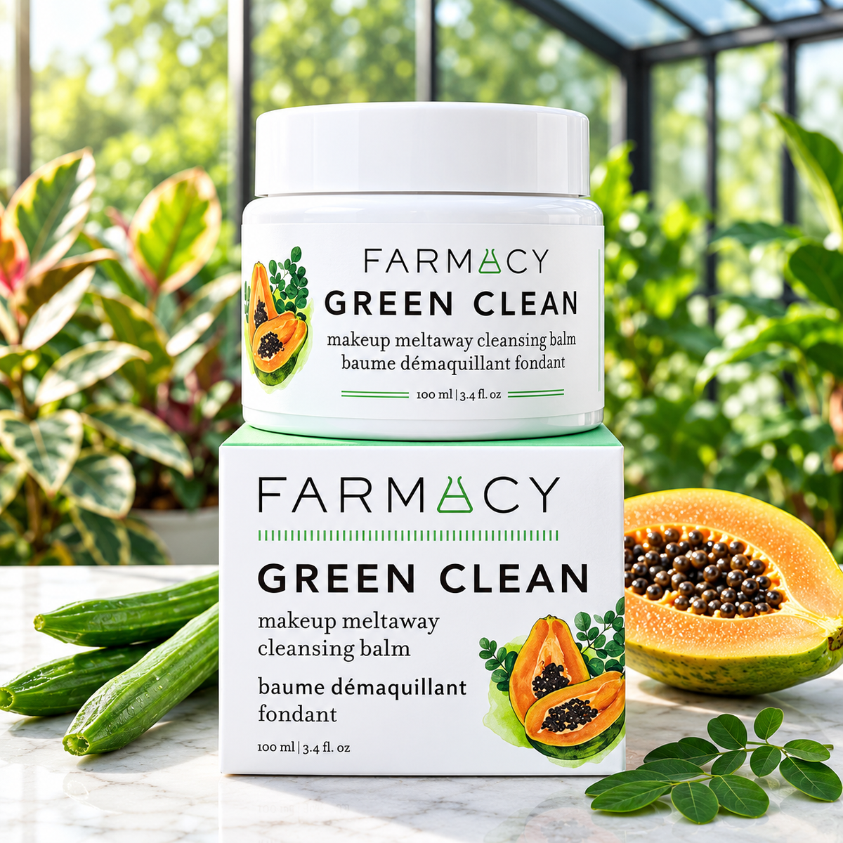 A jar of FARMACY Green Clean makeup meltaway cleansing balm sits on top of its matching box, styled on a marble surface with cucumbers and a sliced papaya in a lush greenhouse setting.